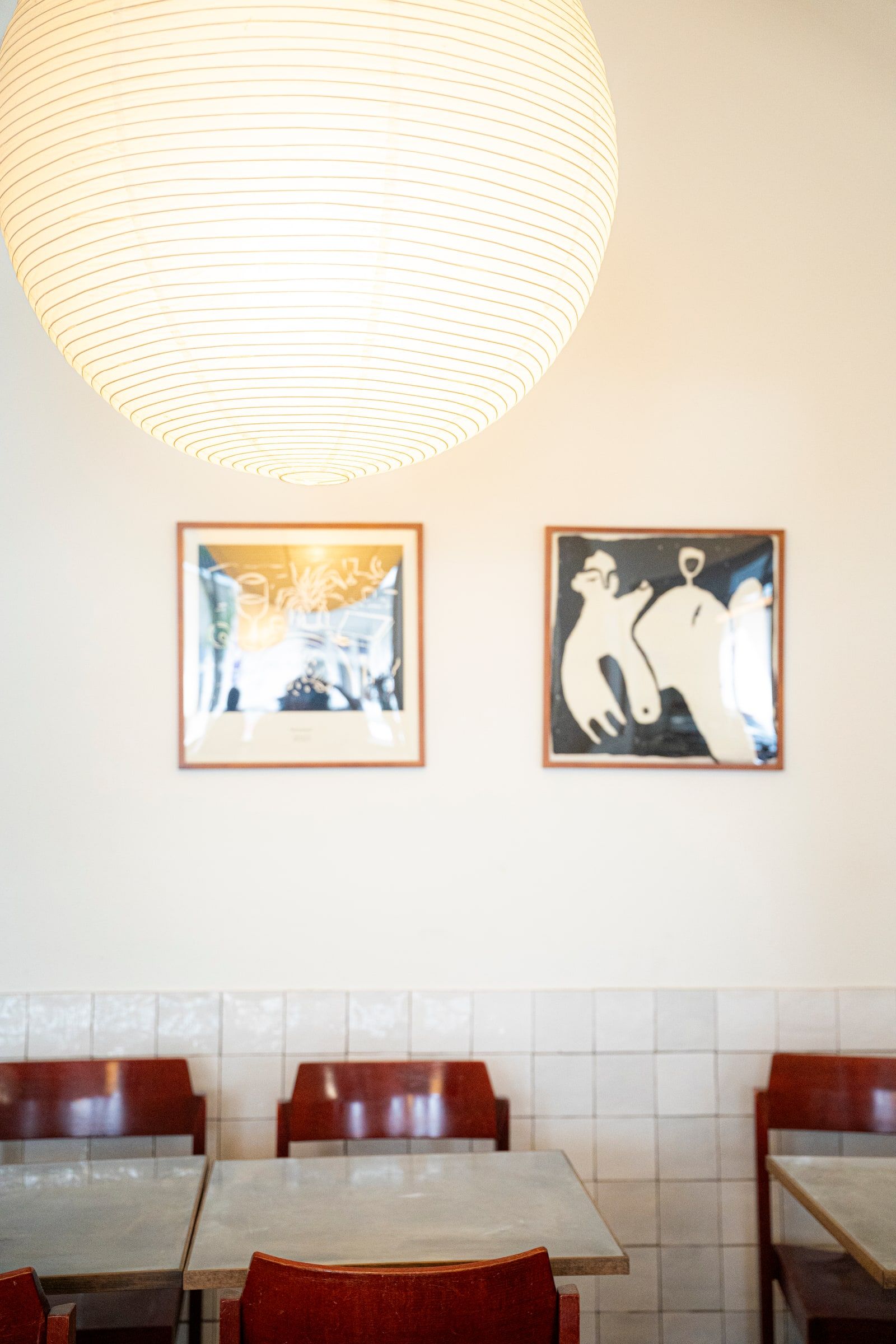 Two framed prints on a cream wall above red chairs, under a paper lantern